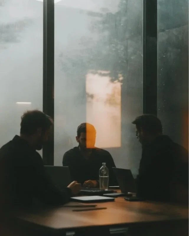 Three people engaged in a discussion in a darkened conference room, with a large window behind them showing a foggy outdoor scene.