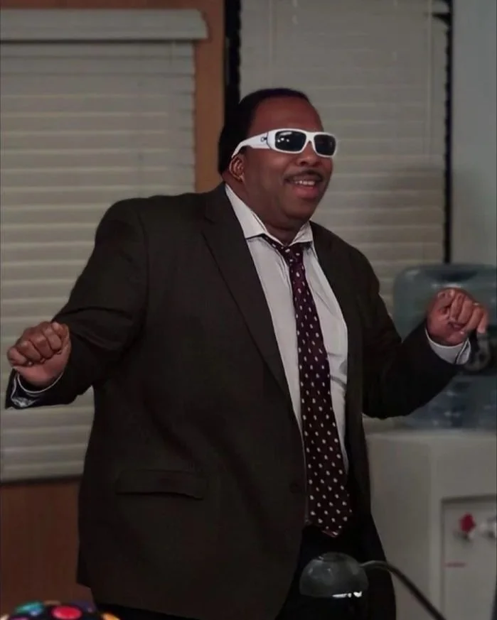 Man in a suit wearing sunglasses, smiling with fists raised, standing indoors near window blinds.