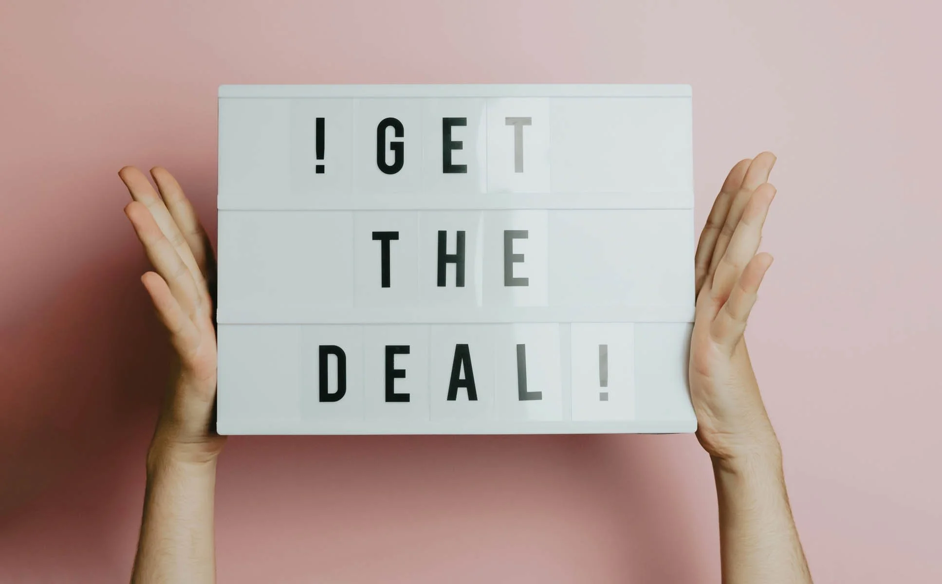 Kaksi kättä pitämässä valkoista valotaulua, jossa lukee '! GET THE DEAL!' vaaleanpunaisella taustalla.