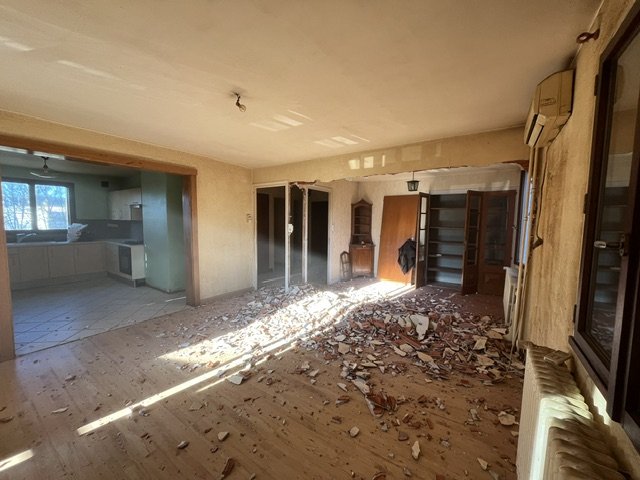 Pièce endommagée avec débris de plâtre au sol, murs en mauvais état, une fenêtre, une porte, une bibliothèque vide, et un chauffage.