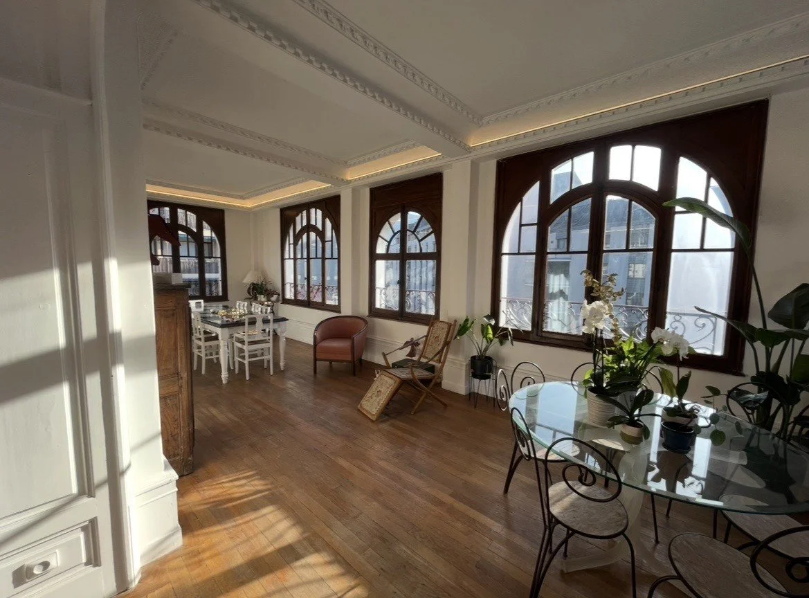 Salon lumineux avec parquet en bois, grandes fenêtres avec cadre en bois, fauteuil en cuir, table à manger ronde avec chaises en fer forgé, plantes en pot et décoration intérieure élégante.