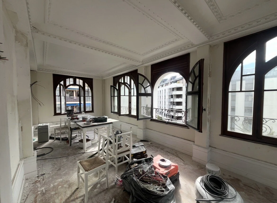 Pièce en rénovation avec de grandes fenêtres en bois arquées, des meubles blancs et des matériaux de construction au sol.