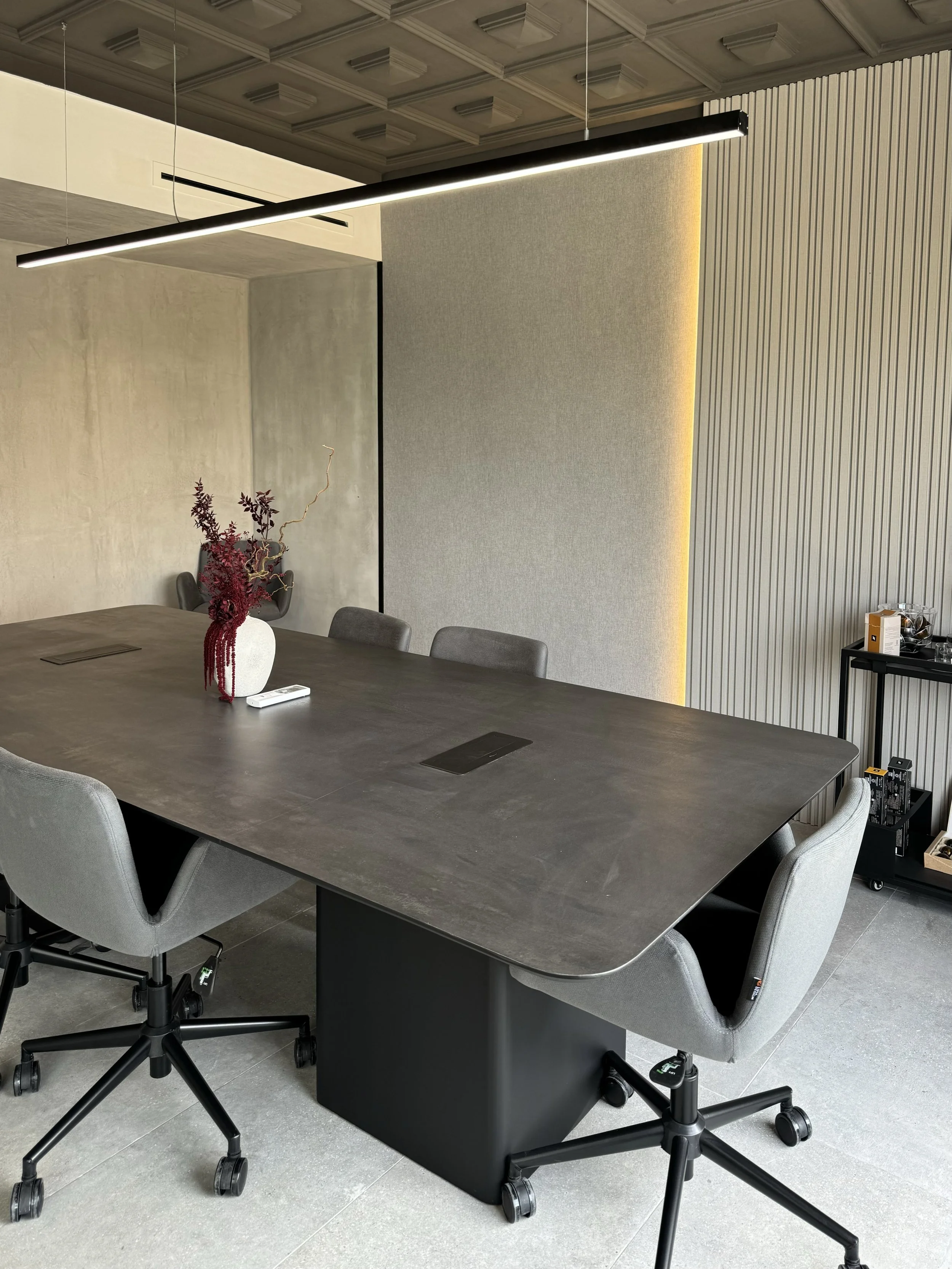 Salle de réunion moderne avec une grande table en bois foncé, six fauteuils gris, une pochette avec une plante rouge dans un vase blanc, et un serveur d'alcool ou de boissons dans un coin. Suspension lumineuse longue suspendue au plafond. Mur avec décoration en panneaux en bois et en tissu. Sol en carrelage gris.