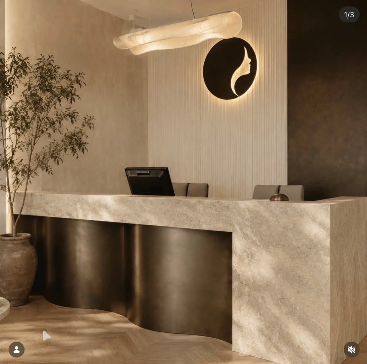 Un comptoir de réception en pierre avec un fond beige et une décoration moderne. Un arbre en pot se trouve à gauche de l'image, et un éclairage suspendu style nuage est au plafond. Une œuvre d'art représentant une lune ou un symbole circulaire est accroché au mur derrière le comptoir.
