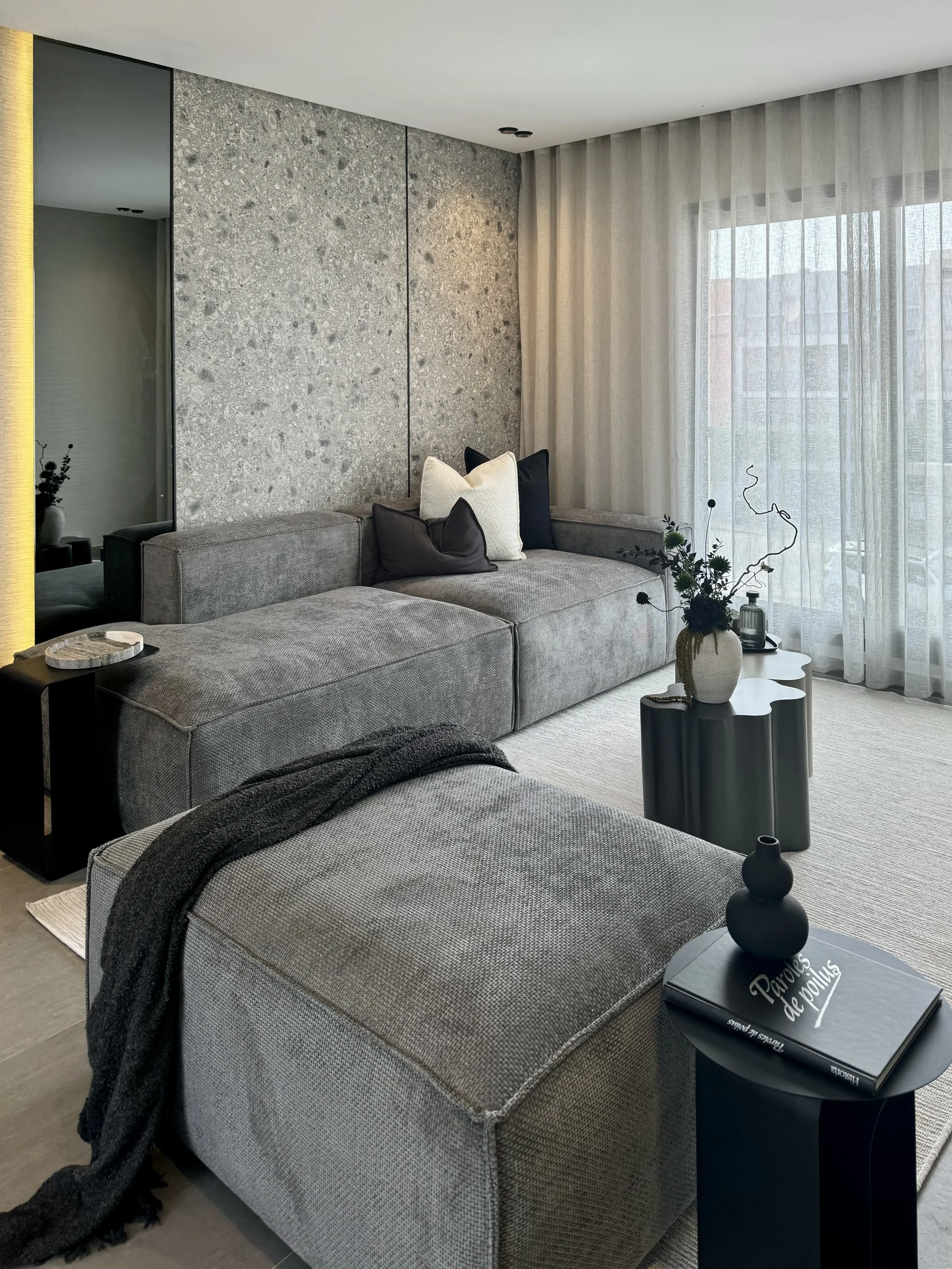 Salon contemporain avec canapé gris, coussins noirs et blancs, table d'appoint, vase avec des branches décoratives, grande fenêtre avec rideaux légers