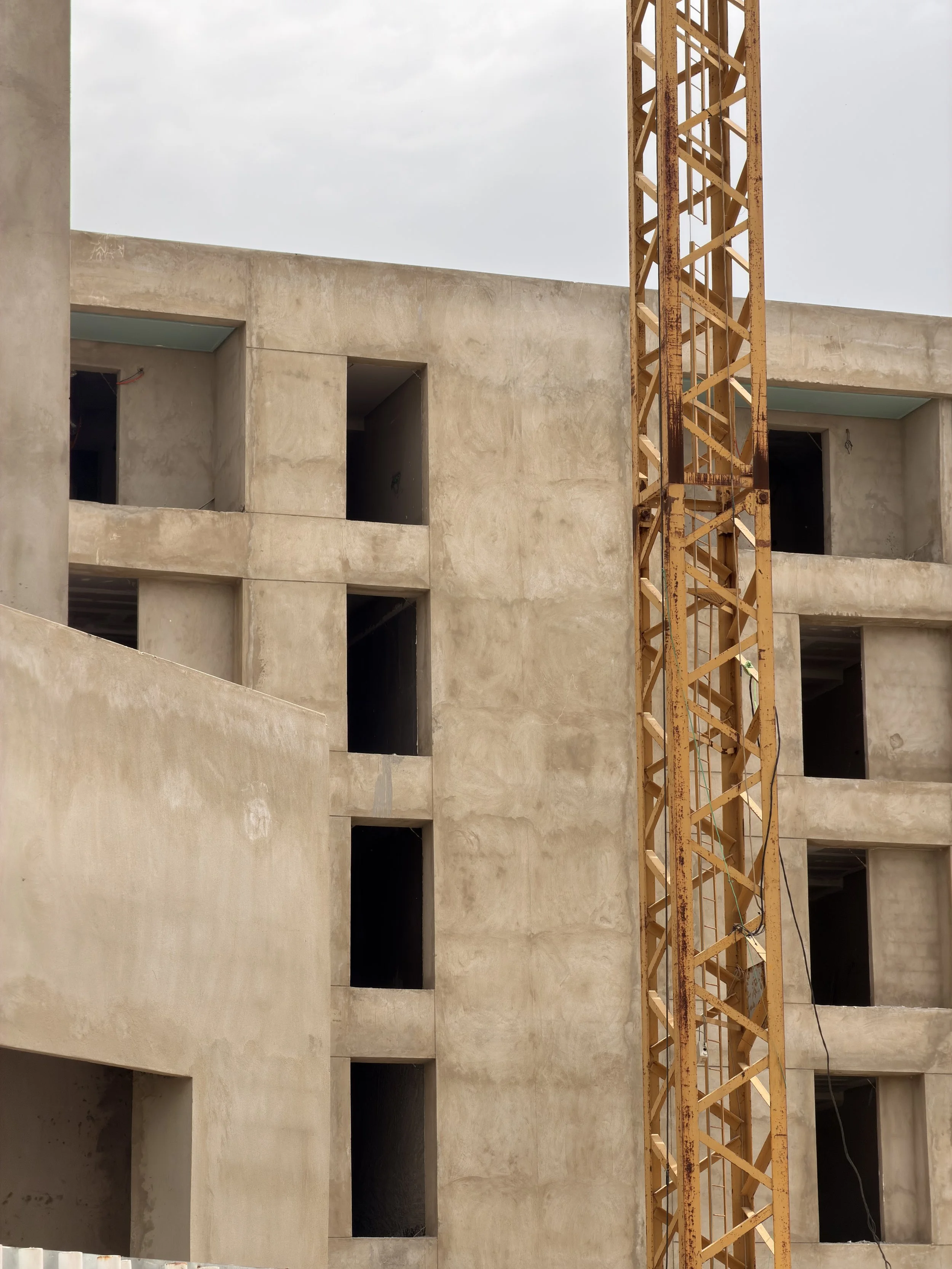 Construction d'un bâtiment en béton avec une grue de chantier orange à côté, fenêtres inachevées, ciel nuageux.