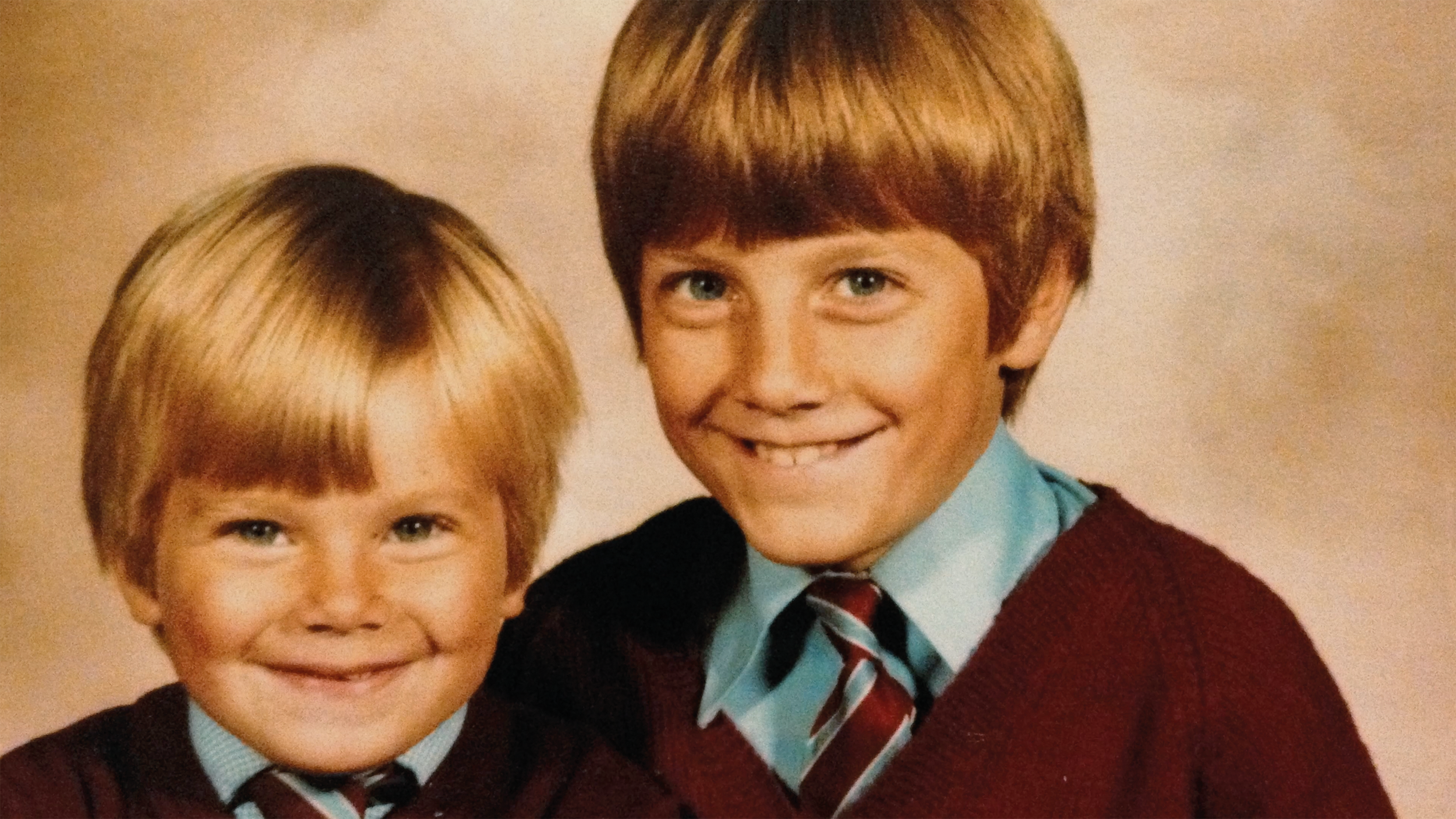 Two young boys in school uniforms smiling, with one boy slightly behind the other, against a plain background.
