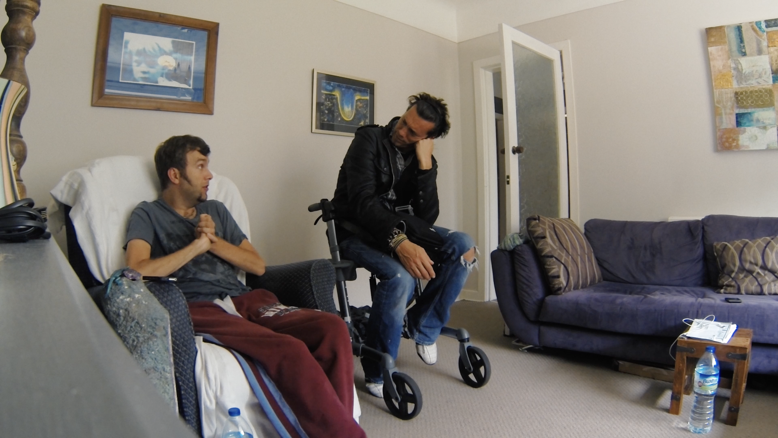 Two men are in a living room, one sitting on a couch with his hands clasped and the other sitting on a wheelchair, leaning forward with his hand on his head, engaging in conversation. The room has framed artwork on the walls, a patterned sofa, a side table, and a water bottle on the floor.