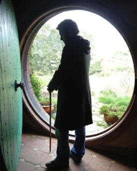 Silhouette of an elderly person with a cane standing in a circular doorway, overlooking a garden with lush greenery.
