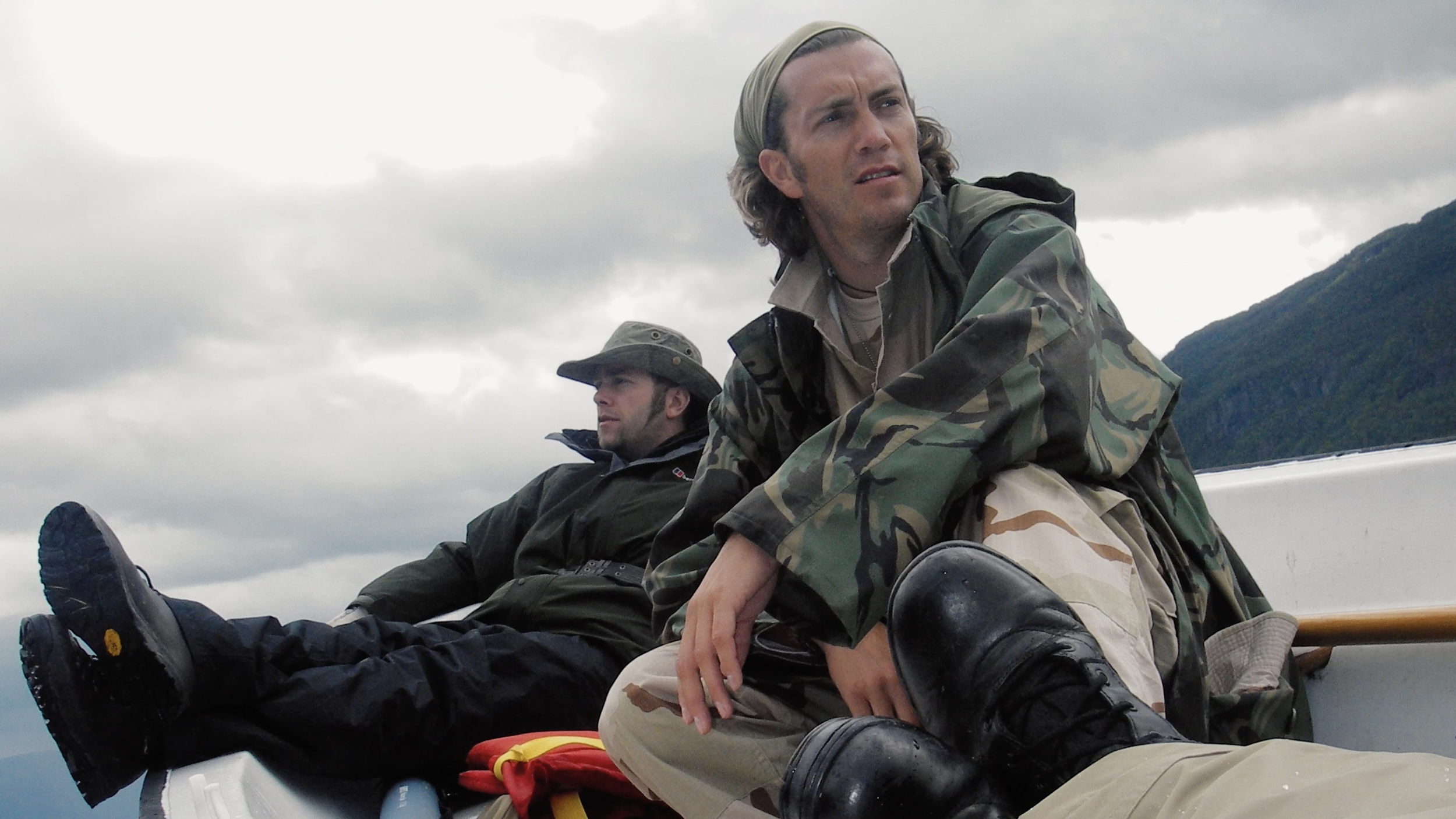 Two men in camouflage clothing and hiking gear sitting on a boat, with cloudy mountain landscape in the background.