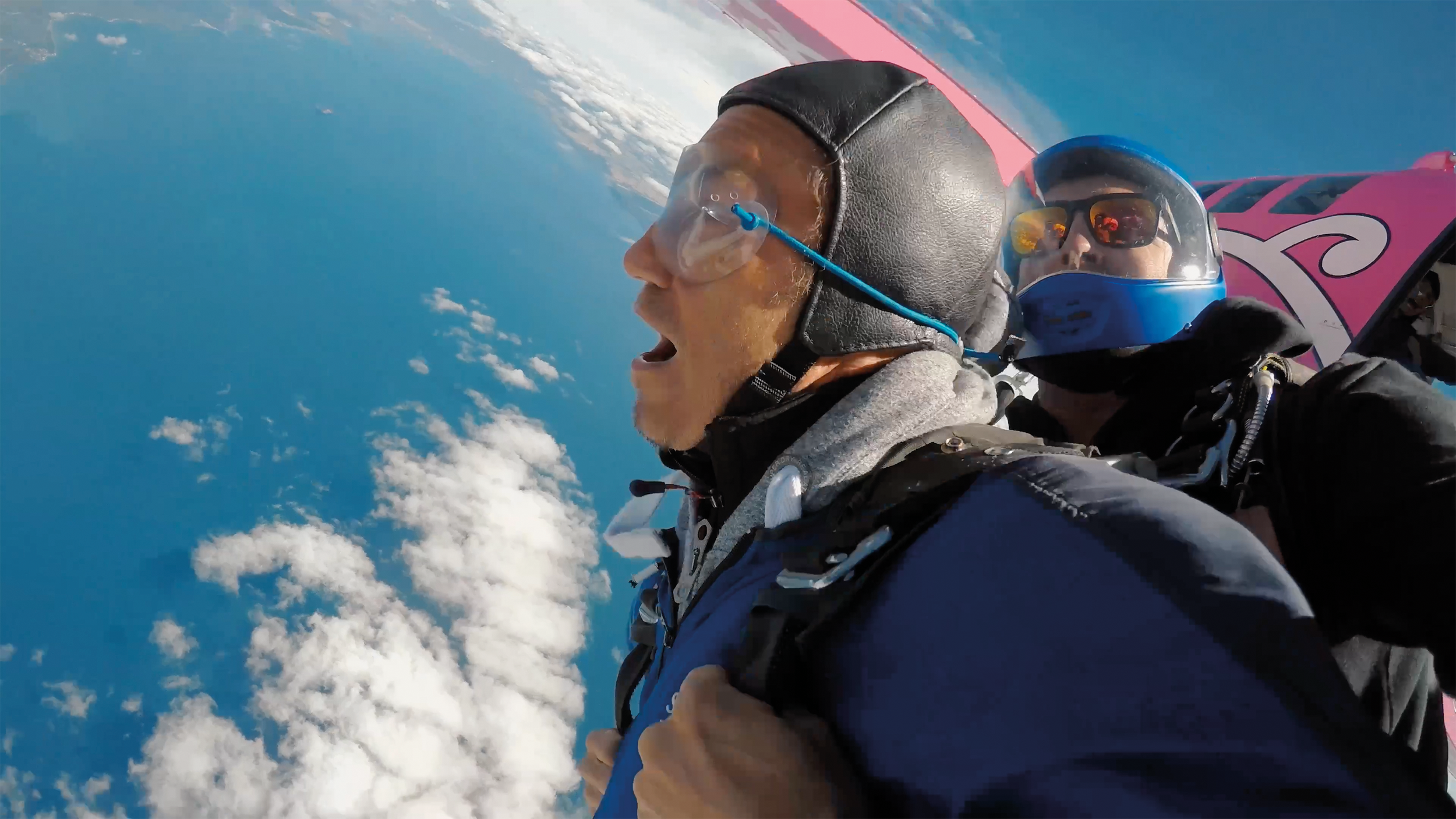 Two skydivers in mid-air, one in a black helmet and goggles with an open mouth, and the other in a blue helmet and sunglasses, with clouds and blue sky in the background.