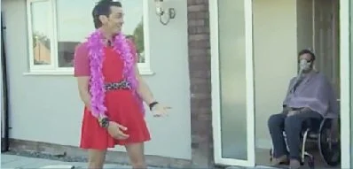 Person dressed in a pink dress with a feather boa, standing outdoors near a house, speaking to a person sitting in a wheelchair.