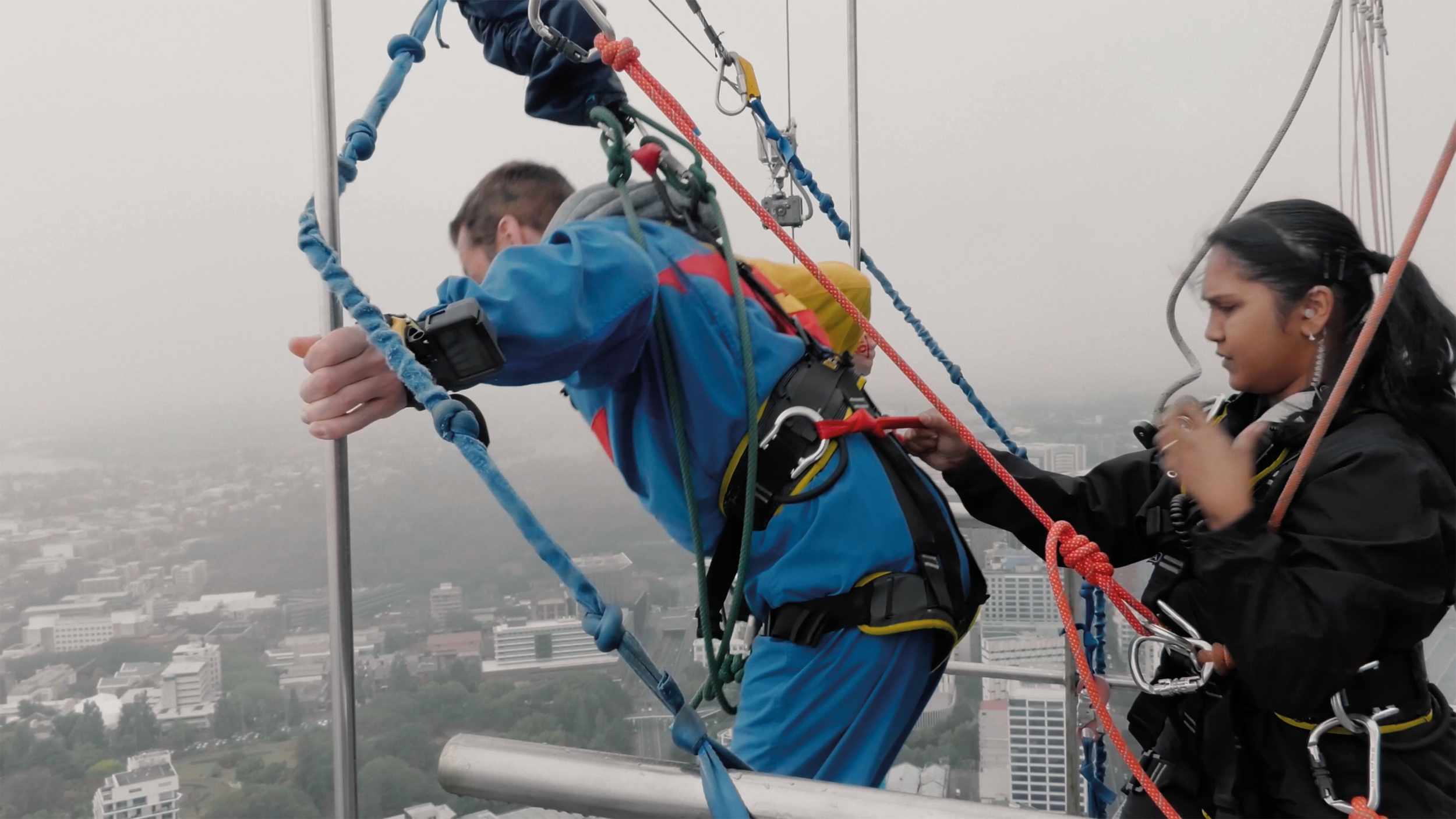 Two people are high above the city on a tall structure, engaged in a construction or maintenance activity. They are using safety harnesses, ropes, and helmets for protection, with scaffolding or rigging surrounding them.