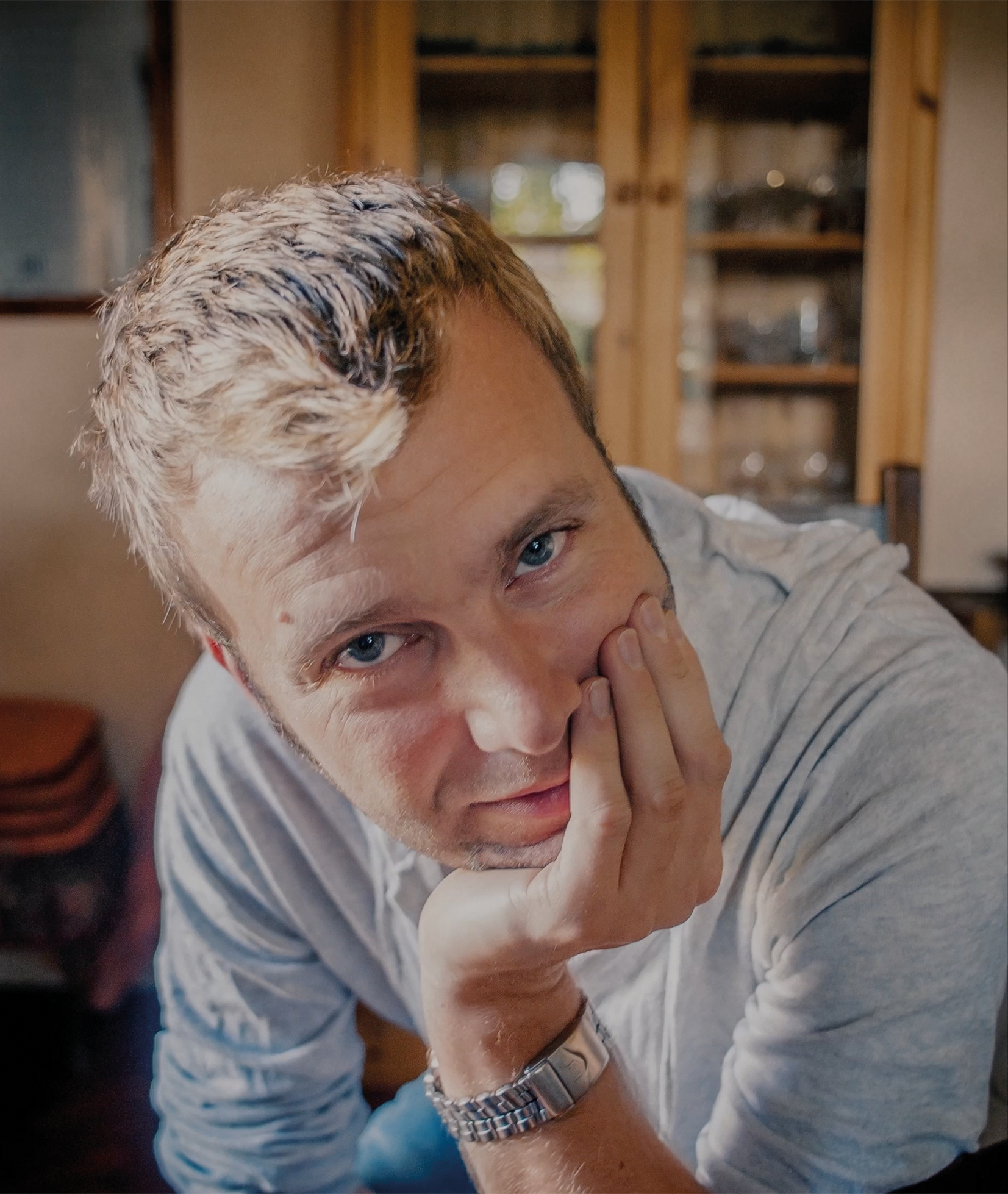 Close-up of a young man with blond hair and blue eyes, resting his face on his hand, wearing a silver wristwatch and a grey striped shirt in an indoor setting.