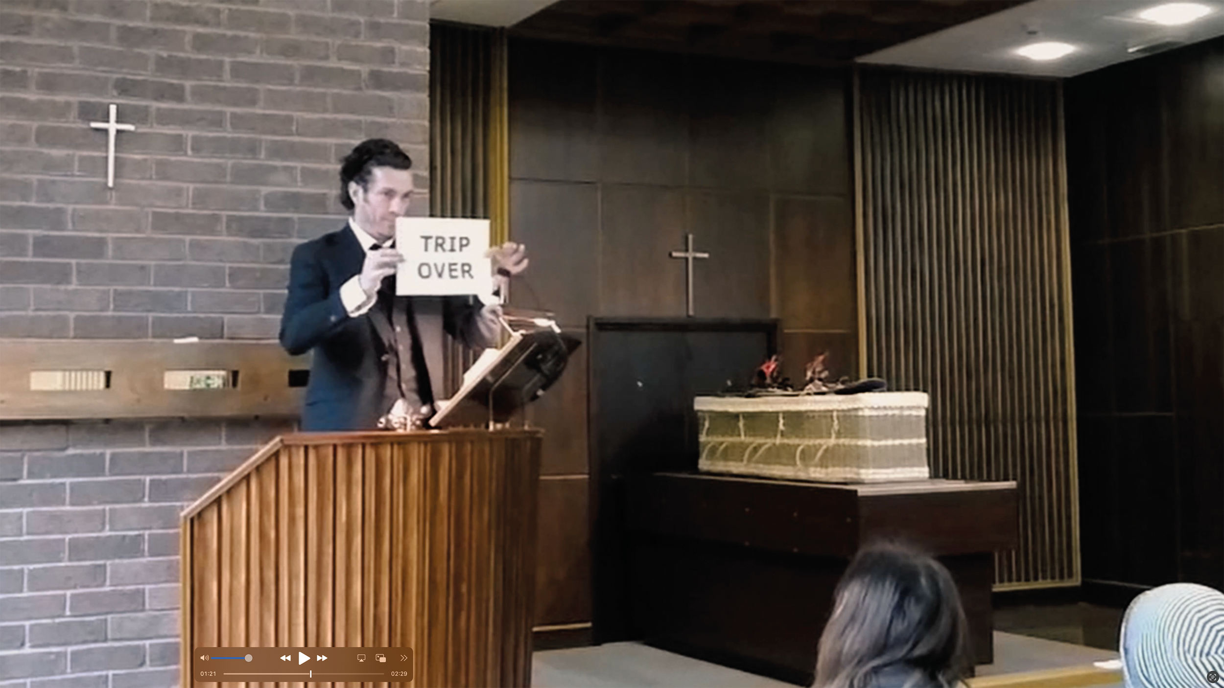 A man dressed in a dark suit and bow tie standing at a church pulpit, holding a sign that reads "TRIP OVER", during a church service or event. The church interior features brick walls, crosses, and a piano.