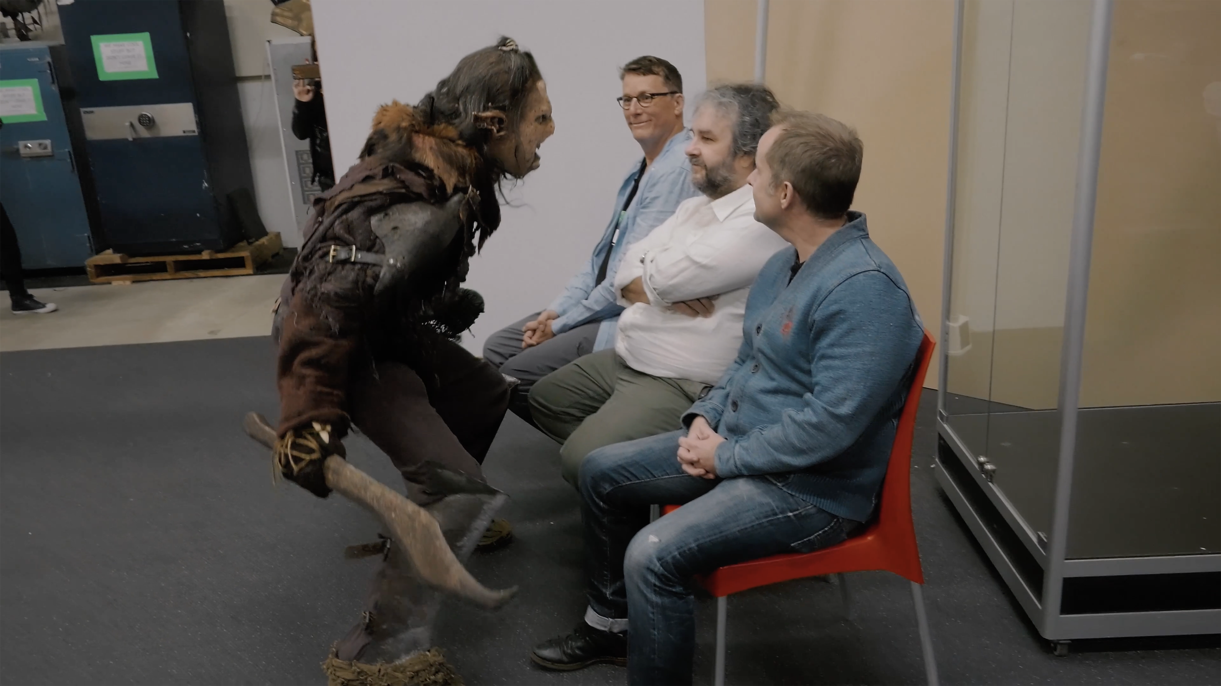A person dressed as a fantasy creature confronts three seated men smiling and watching. The scene appears to take place indoors in a casual setting.