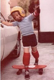 Young child with a helmet balancing on a skateboard, arms outstretched for balance, in a driveway with a car nearby.