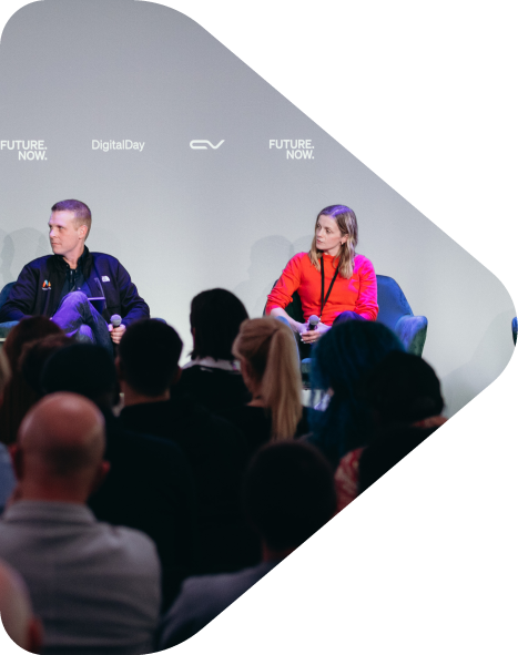 Two people sitting on a stage panel with an audience watching. The background displays the words 'FUTURE. NOW.', 'DigitalDay', and a logo.