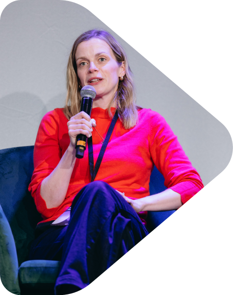 Founder of Flint & Arrow, Kit Powney, sitting on a green chair, holding a microphone, wearing a red top and dark pants, speaking at an event.