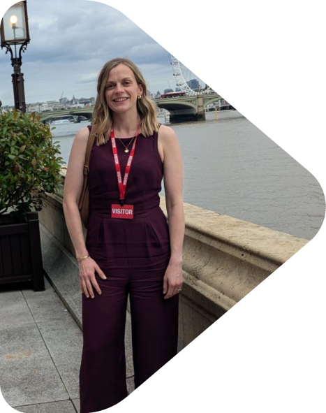 Founder of Flint & Arrow, Kit Powney, stands near a river with city buildings and a bridge in the background, wearing a burgundy sleeveless jumpsuit and a visitor badge.