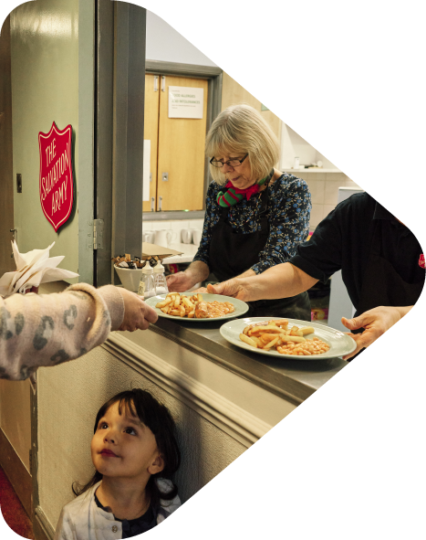 A woman with glasses and a woman in a black shirt serve food to a child at a cafeteria counter, with a young girl gazing up at them.