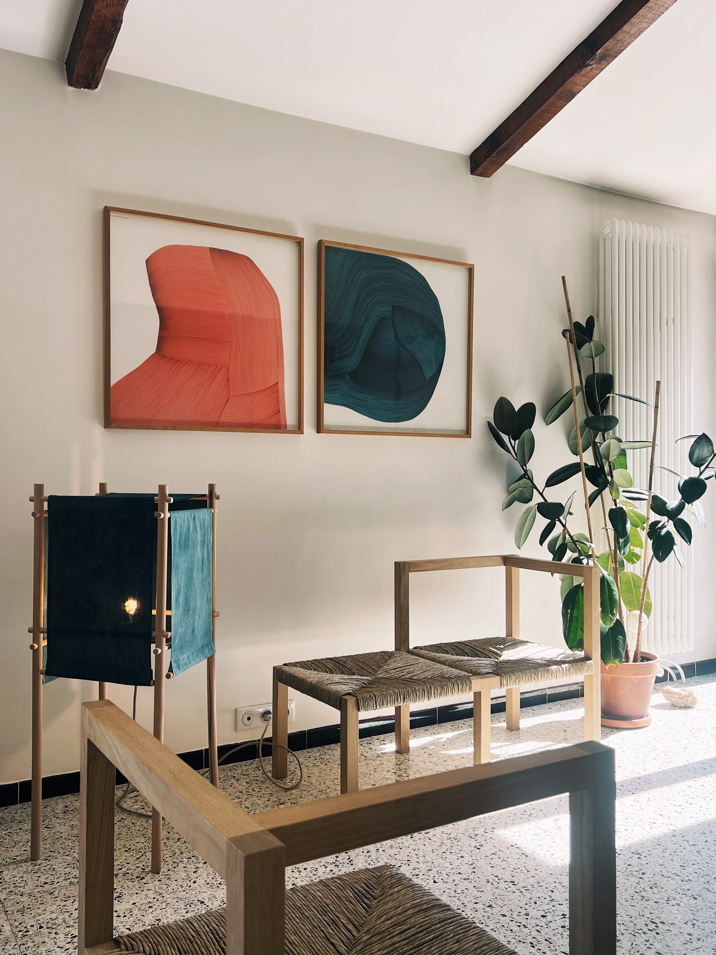 Intérieur minimaliste présentant les créations du designer Alexandre Reignier, Angle, un fauteuil en chêne et assise en paille. Ainsi qu'un lampe sur pied minimaliste en hêtre tamisé par des pans de tissus anciens teintés indigo.