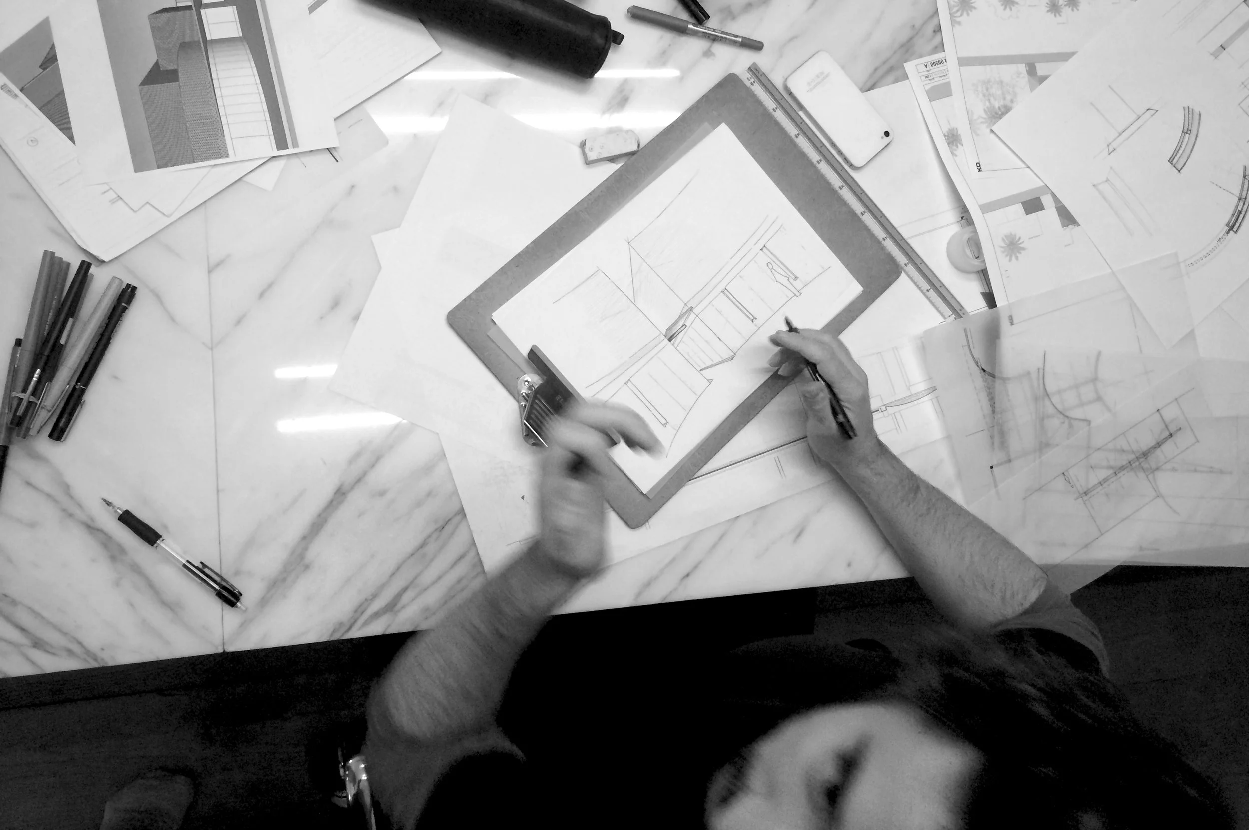 Vue plongeante en noir et blanc d’un architecte dessinant des plans sur une table couverte de croquis, règles et stylos.