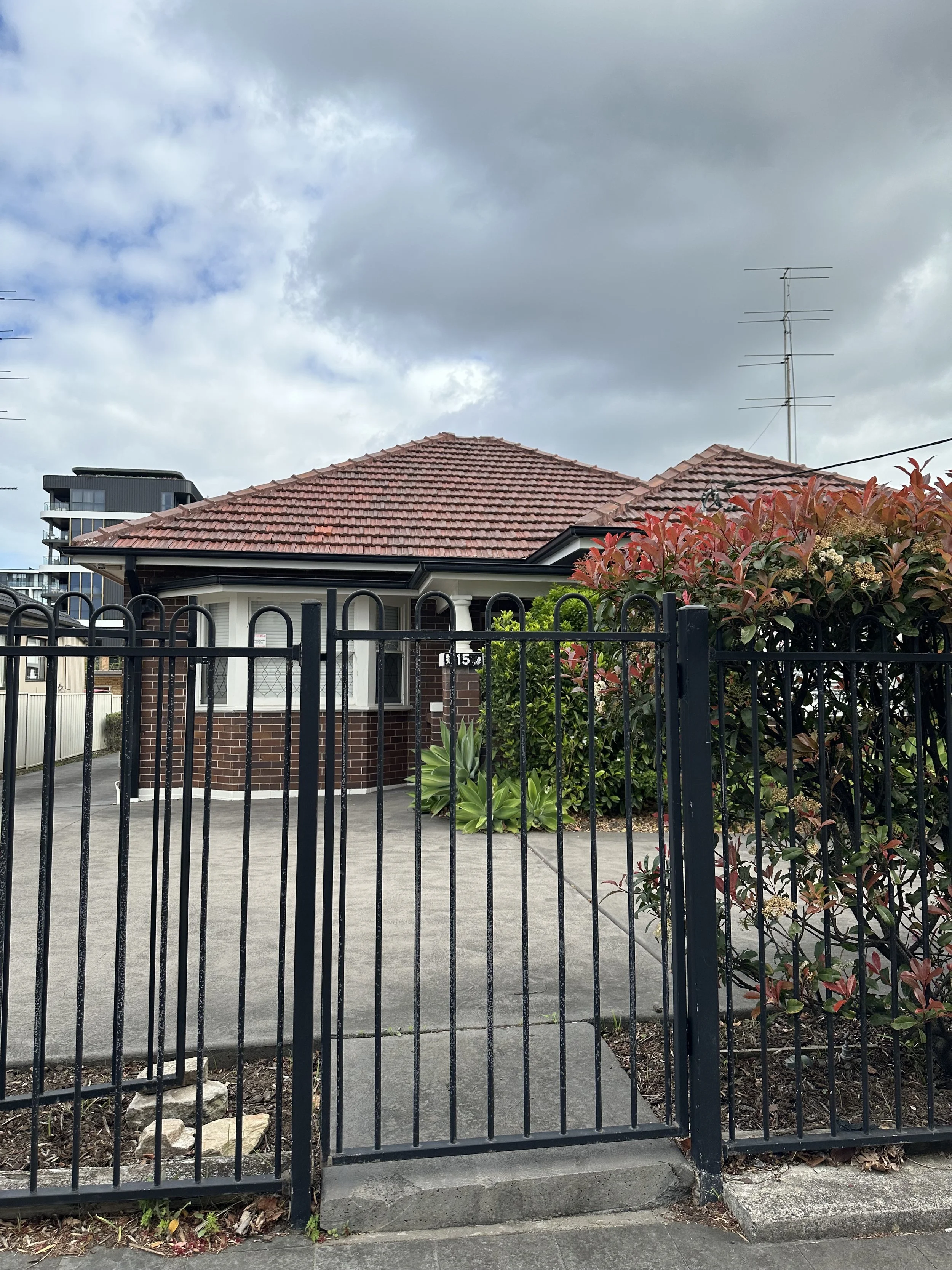 View of a brick house with a red-tiled roof, a black metal gate in the foreground, landscaped garden with plants and shrubs, and an overcast sky with clouds.