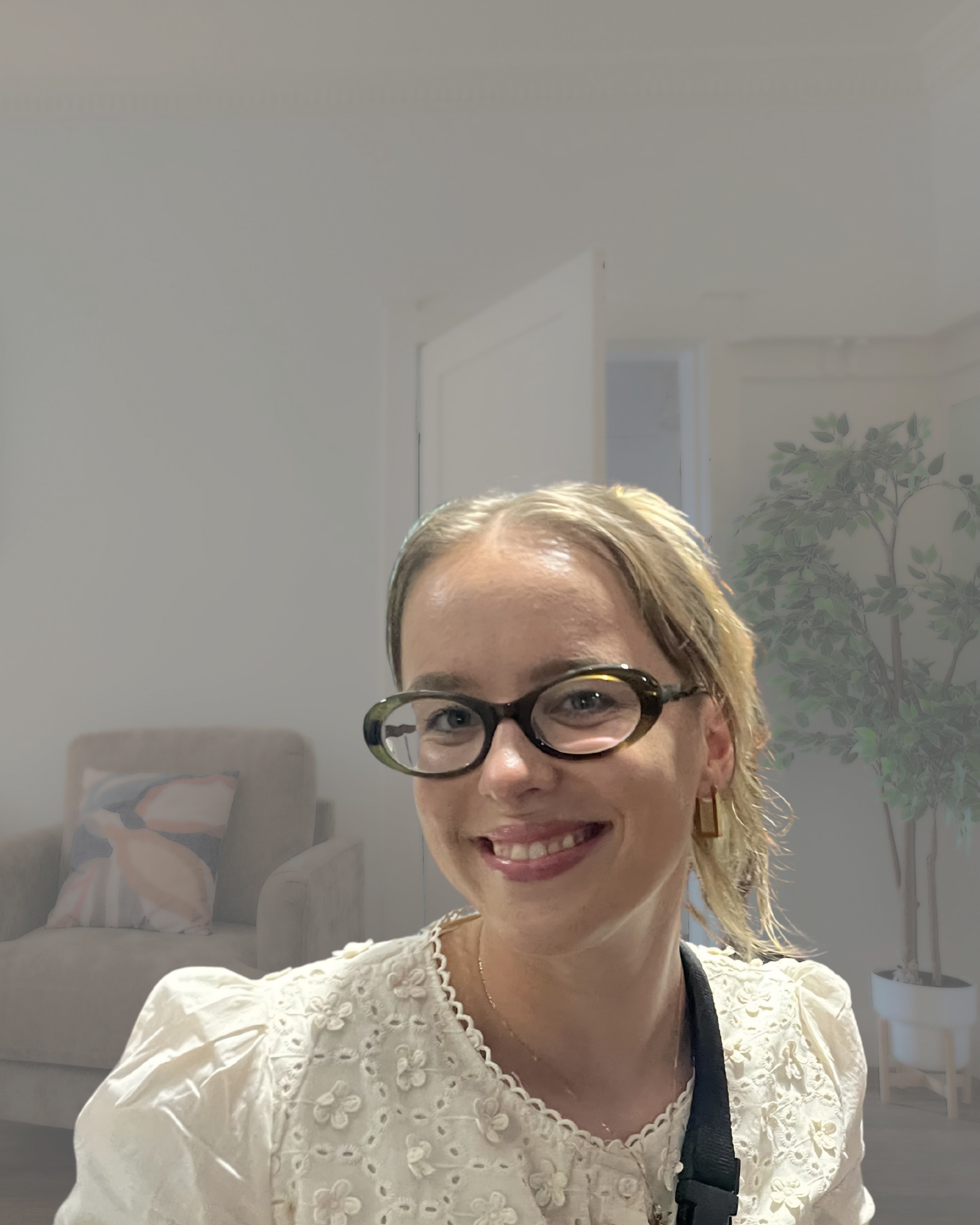A smiling woman with blonde hair, wearing glasses, a white embroidered blouse, and earrings, taking a selfie in a room with a bed, a pillow, a potted plant, and a door in the background.