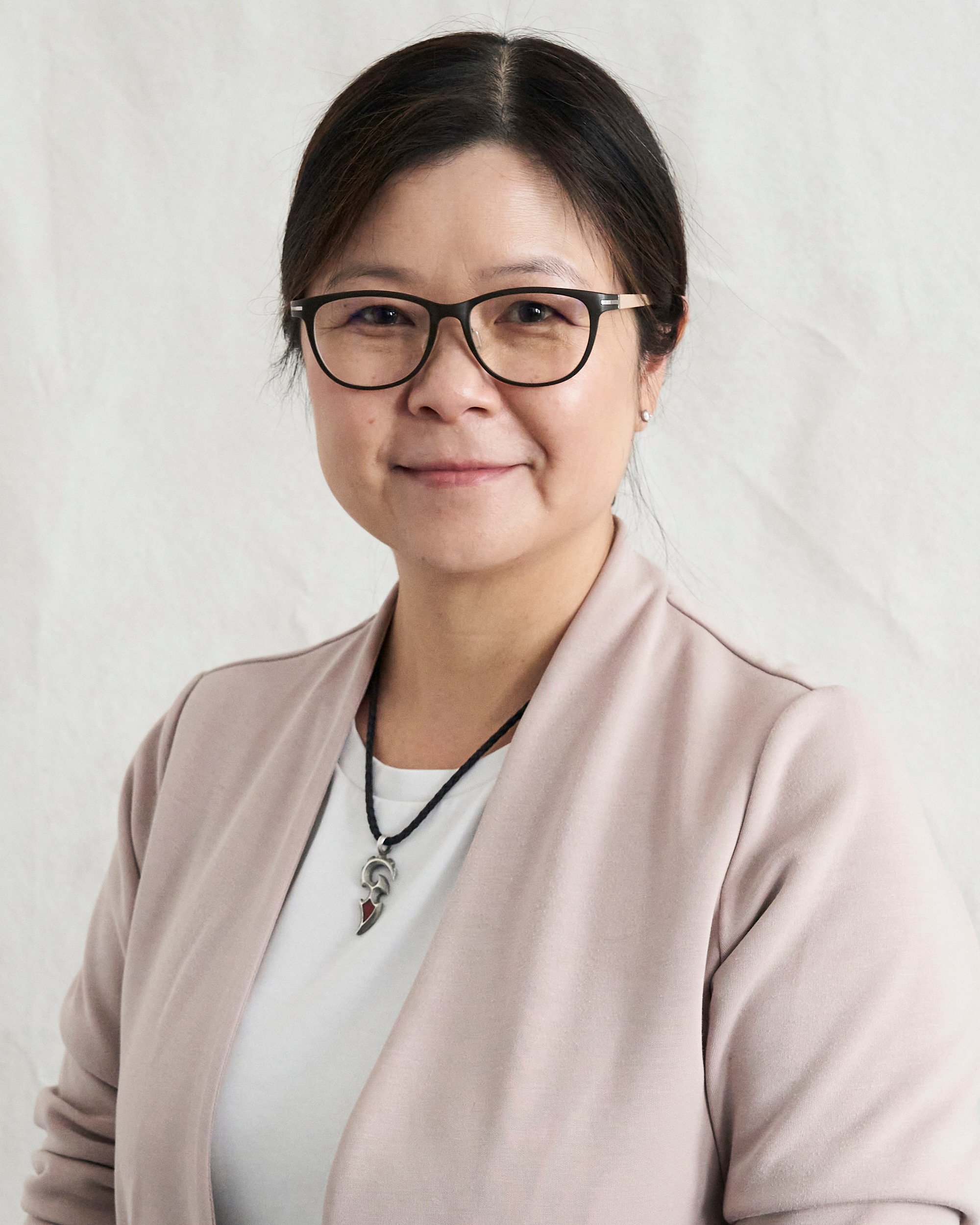 Close-up of a woman with short dark hair wearing black glasses, a beige blazer, and a white top, standing against a plain white wall.