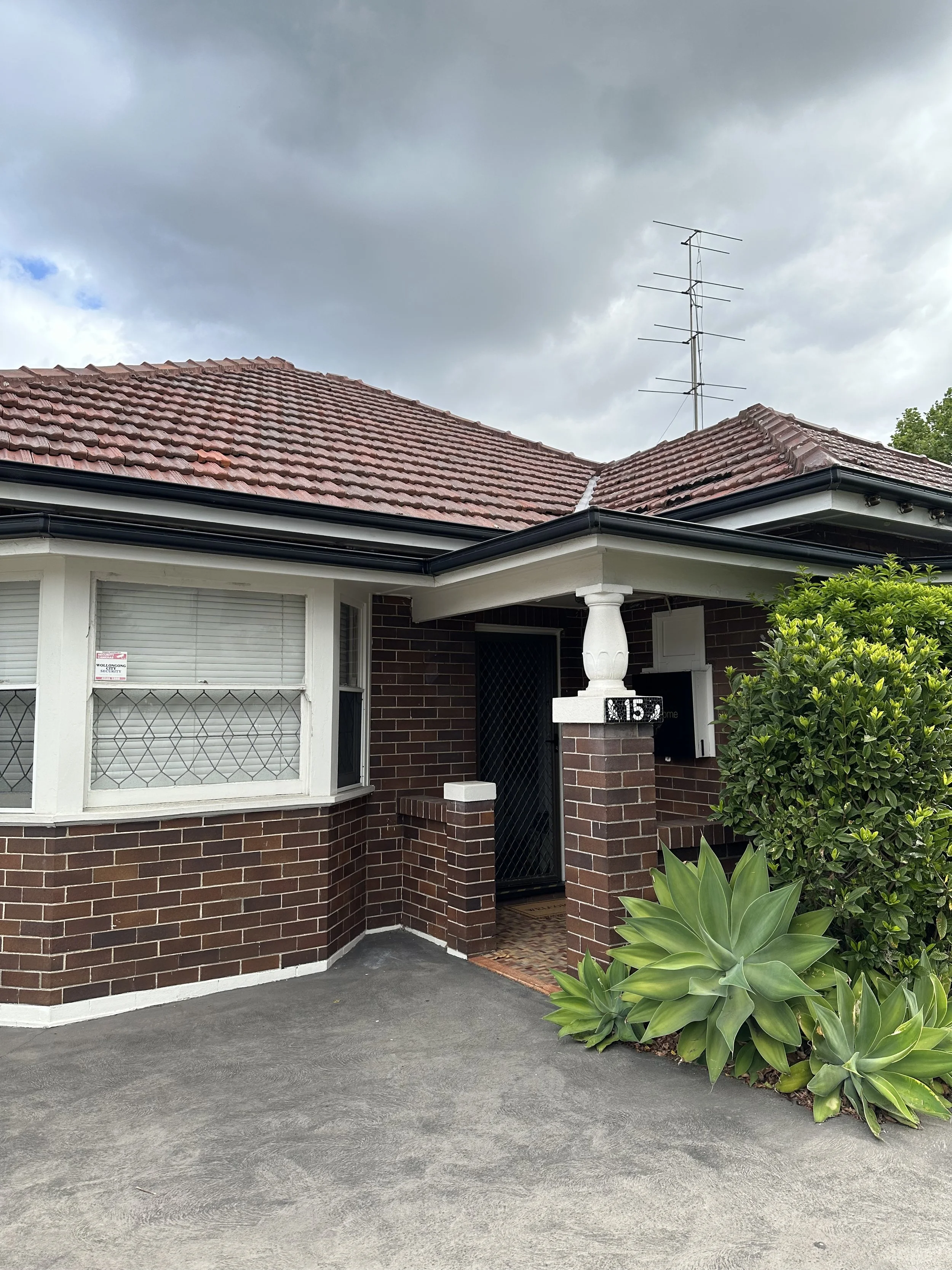 Front view of a brick house with a red tiled roof, a black antenna on the roof, a white window with blinds, a white column supporting the porch, and green plants in front of the house.