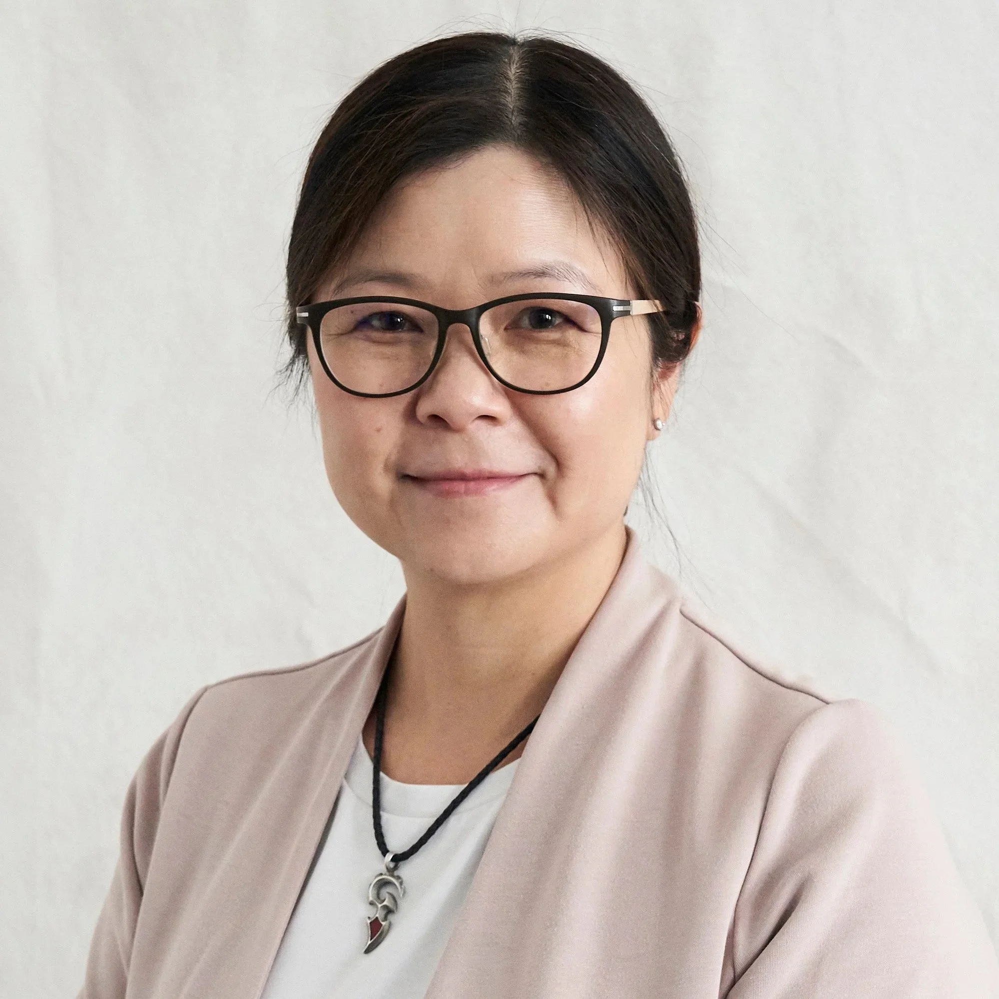Close-up of a woman with short dark hair wearing black-rimmed glasses, a light pink blazer, a white top, a black necklace, and a silver pendant, smiling slightly against a plain light background.