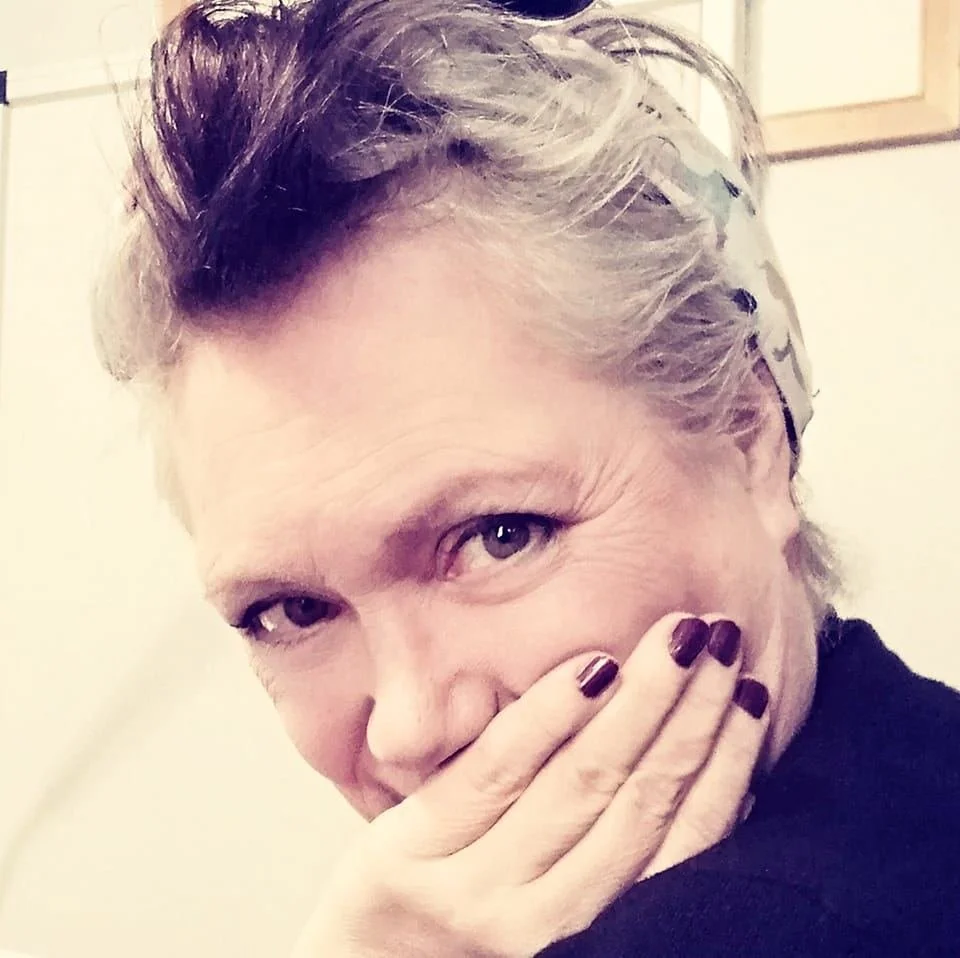 Jools, with short, styled hair covering her mouth with her hand, looking at the viewer, in front of a light-colored wall.