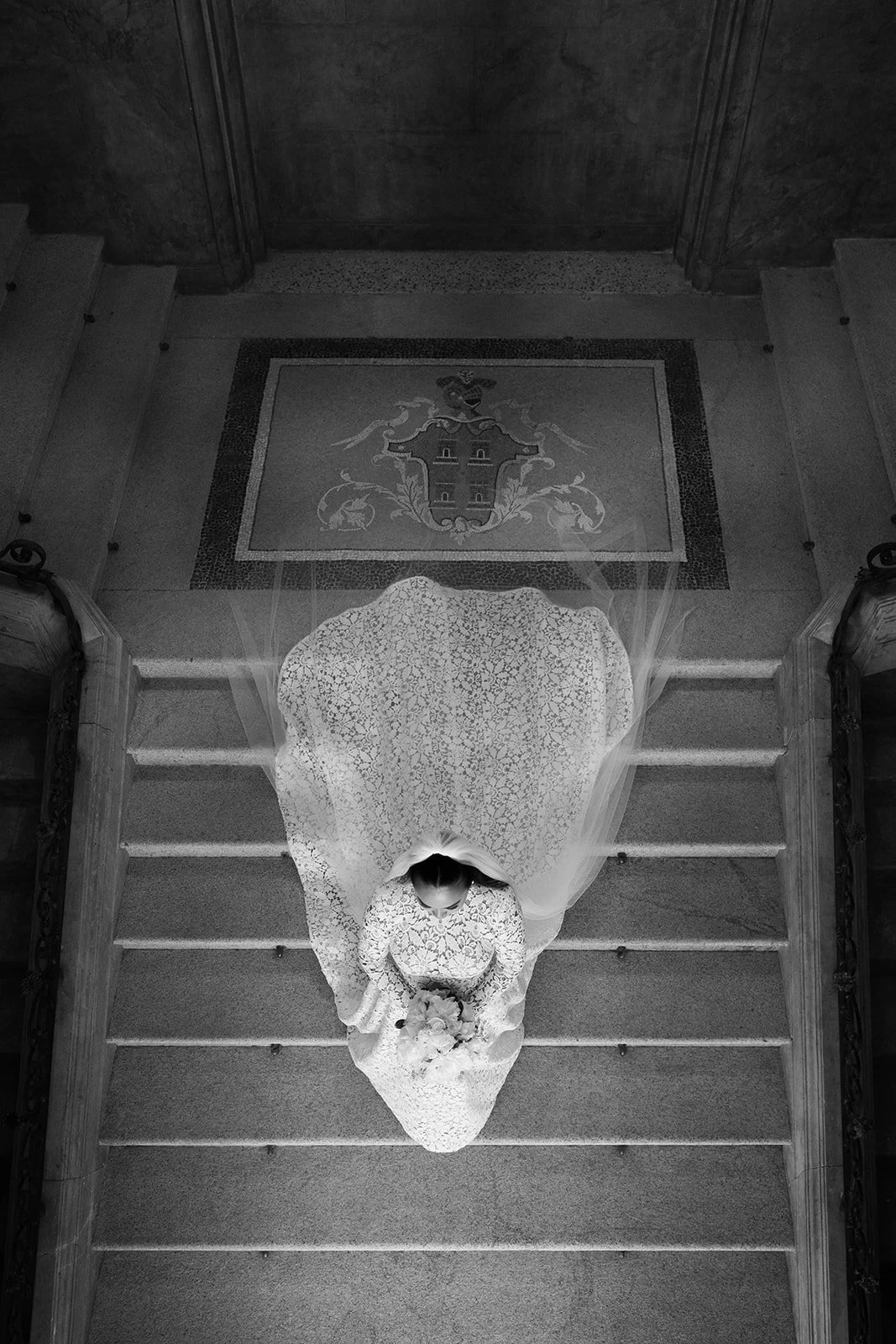 Eine Braut in einem weißen Hochzeitskleid steht vor einer Treppe in einem historischen Gebäude, die vom oberen Blickwinkel aufgenommen wurde.