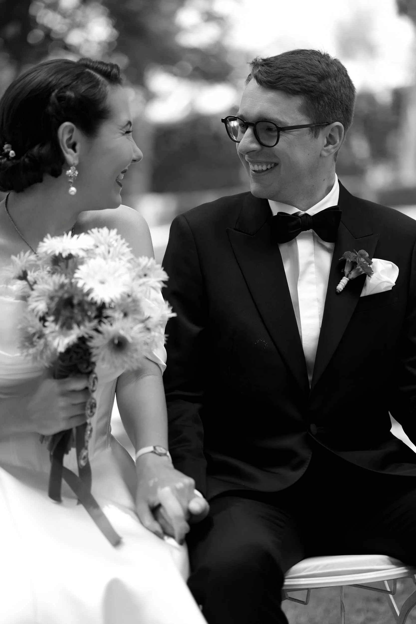 Ein lachendes Paar bei einer Hochzeit, der Mann trägt einen schwarzen Smoking mit Fliege, die Frau trägt ein Brautkleid und extravaganten Ohrringe, beide halten sich an den Händen, im Hintergrund unscharf mit Bäumen.