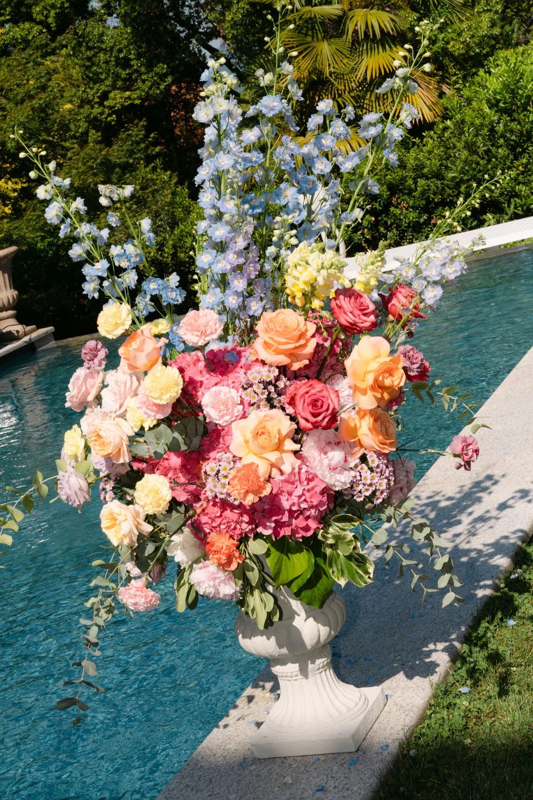 Bunter Blumenstrauß mit Rosen, Hortensien und anderen Blumen in einer weißen Vase neben einem Pool auf einer Steinterrasse, umgeben von grünen Büschen und Bäumen.