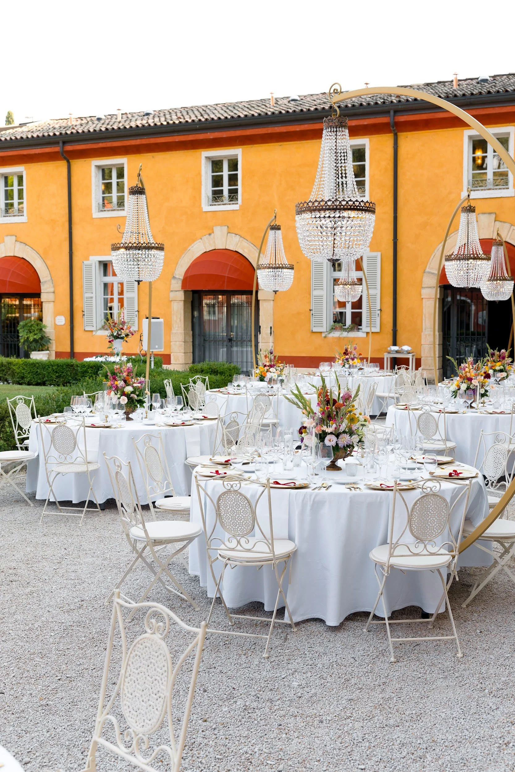 Festlich gedeckte Tische mit weißen Tischtüchern, Gläsern, Besteck und Blumenarrangements vor einem orangefarbenen Haus mit weißen Fenstern und Dachrinnen, verziert mit großen Kristallkronleuchtern.