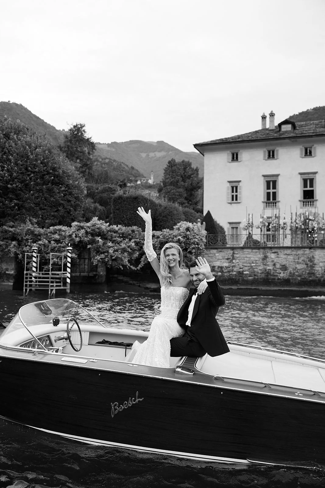 Ein frisch vermähltes Paar auf einem Boot im Wasser, lächelnd und winkend, umgeben von einer malerischen ländlichen Kulisse mit Häusern und Hügeln im Hintergrund