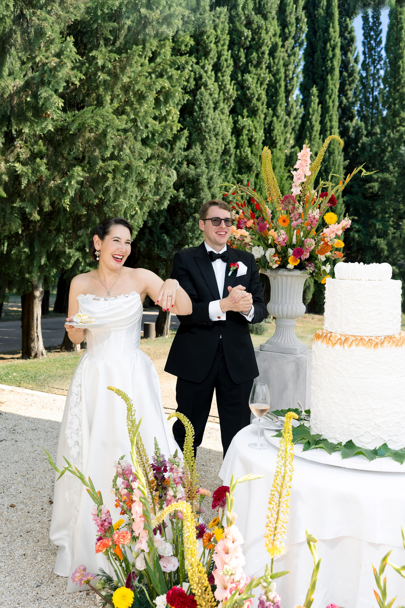 Ein Hochzeitsfest im Freien mit einem lachenden Brautpaar, einer großen Blumenarrangement und einem weißen Hochzeitstorte bei Sonnenschein.