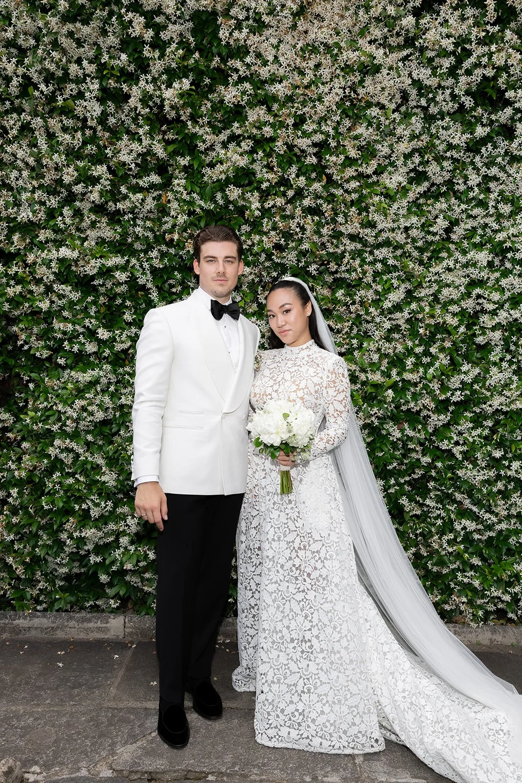 Ein Brautpaar bei seiner Hochzeit, stehend vor einer grünen Wand voller weißer Blumen. Der Mann trägt einen weißen Anzug mit schwarzer Fliege, die Frau trägt ein langes, elegantes weißes Spitzenkleid mit Schleier und hält einen Bouquet aus weißen Blumen.