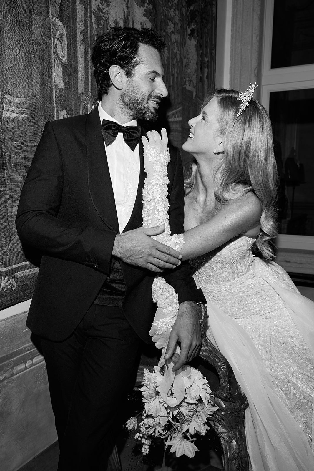 Braut und Bräutigam bei ihrer Hochzeit, lachen und schauen sich liebevoll an, er trägt einen schwarzen Anzug mit Fliege, sie trägt ein weißes Brautkleid mit Schleier und Diadem.
