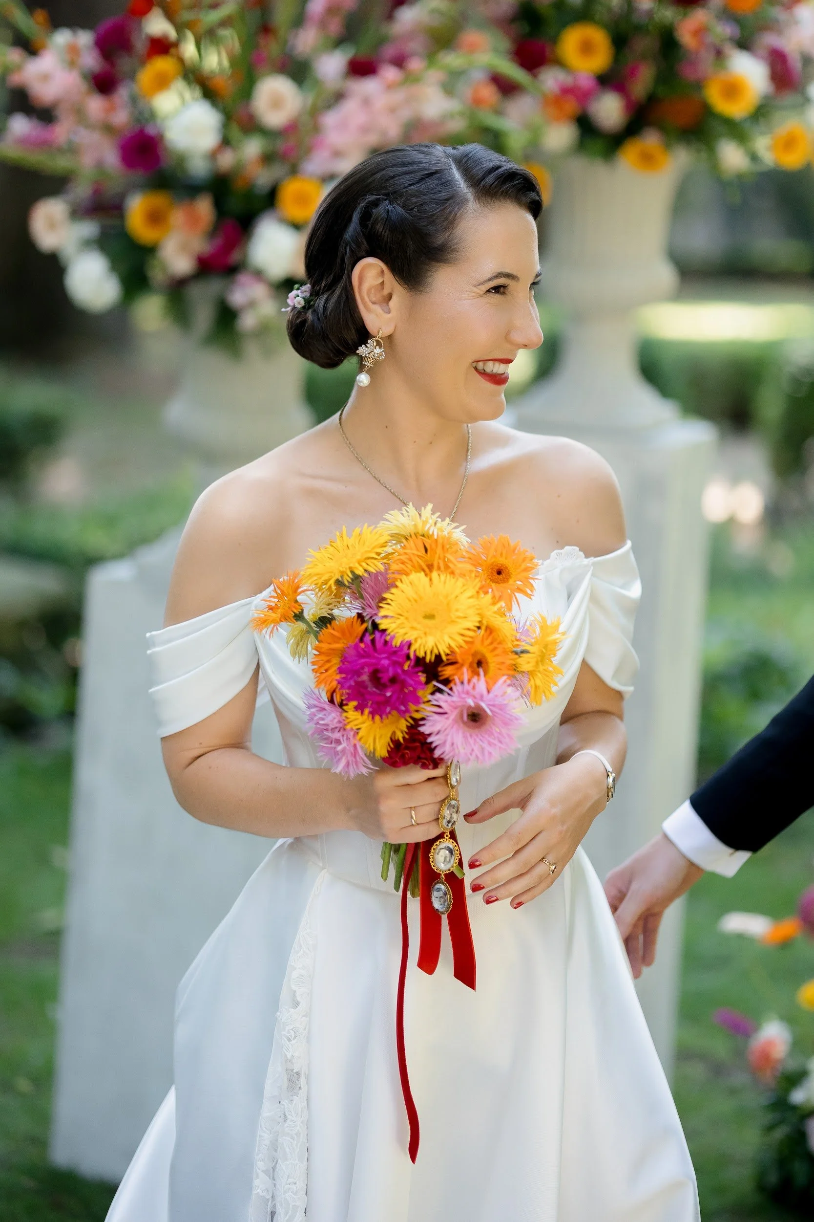Frau in Hochzeitskleid, die einen bunten Blumenstrauß hält, lächelt außerhalb, im Hintergrund sind große Blumenvasen mit bunten Blumen.