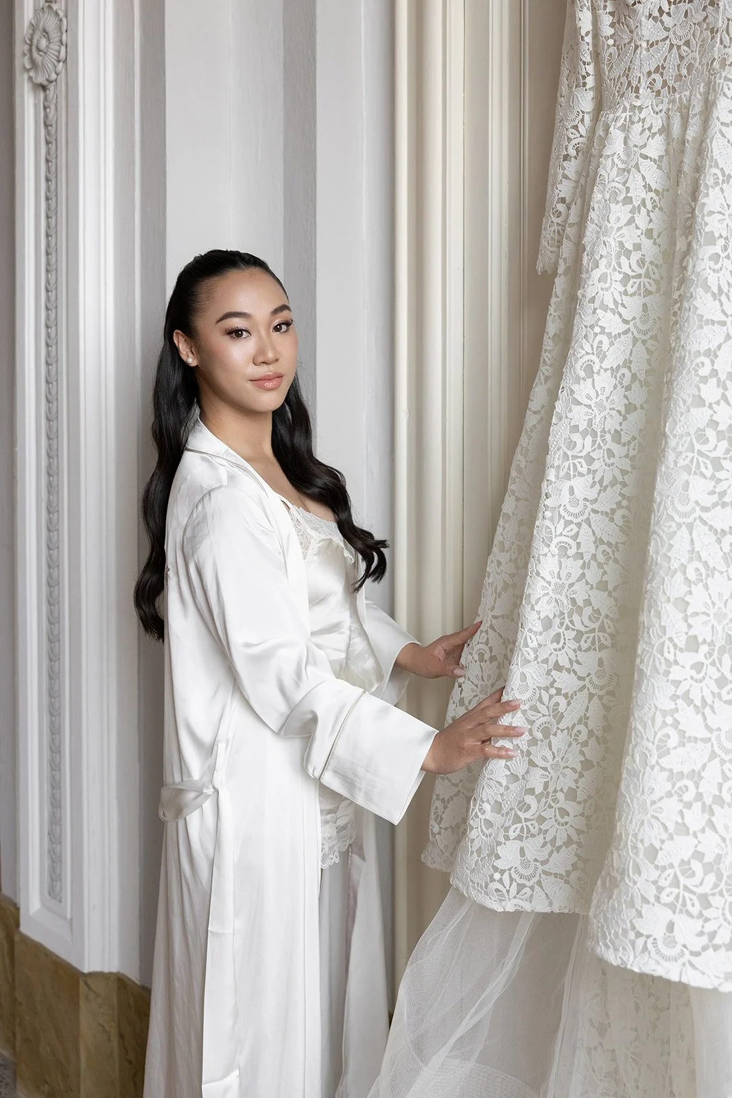 Eine junge Frau in weißem Nachthemd und Satin-Kleid steht neben einer Gardine mit Blumenmuster, die sie vorsichtig berührt.