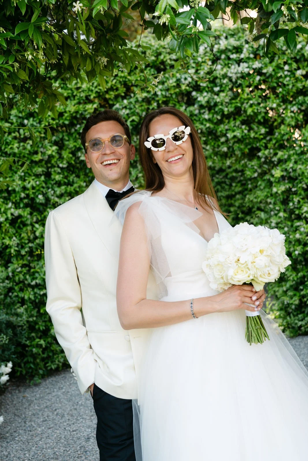 Ein lächelndes Brautpaar bei einer Hochzeit, die Braut trägt eine weiße Hochzeitskleid und hält einen Blumenstrauß, der Bräutigam trägt einen weißen Anzug mit schwarzen Hosen und eine schwarze Fliege, beide tragen lustige Sonnenbrillen, im Hintergrun