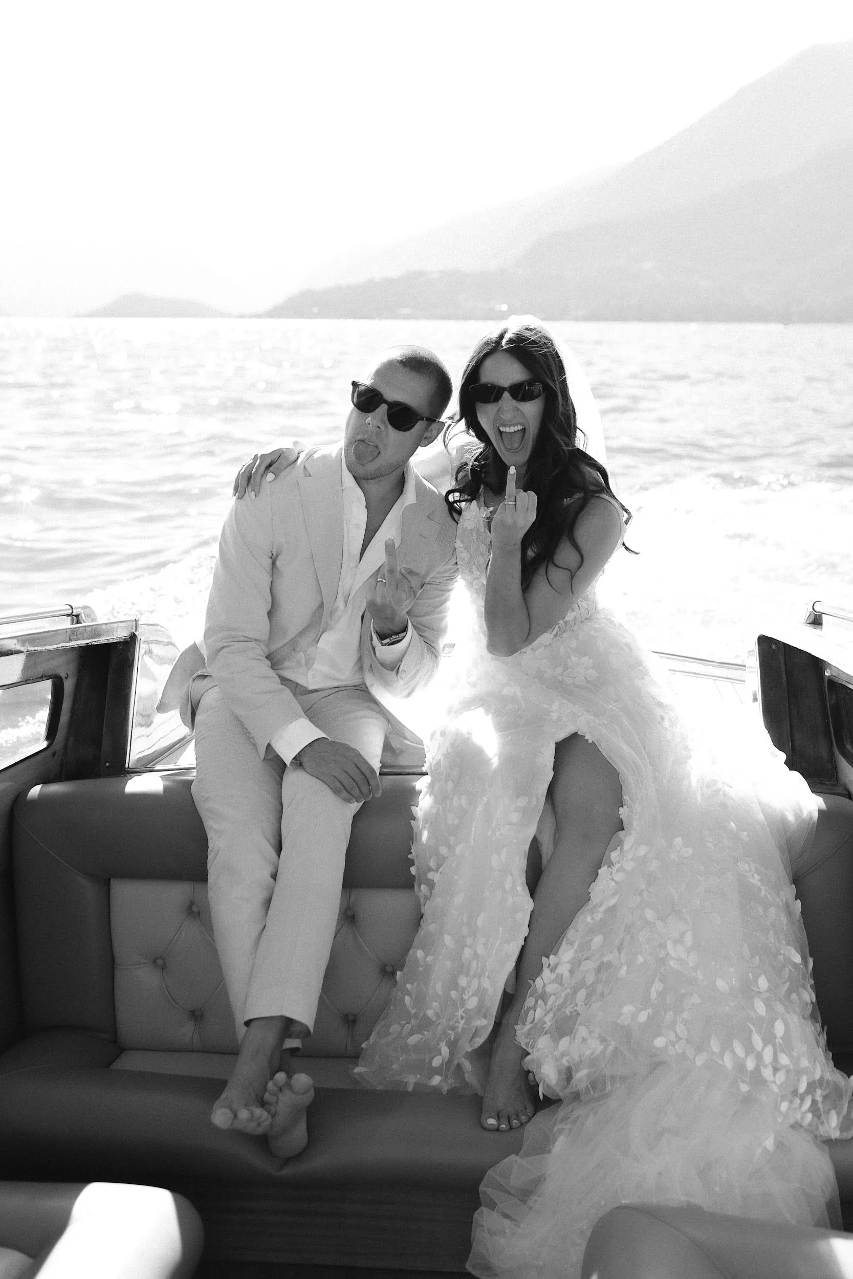 Ein Paar auf einem Boot, vermutlich bei einer Hochzeit, machen Grimassen und zeigen Mittelfinger, während sie im Wasser sitzen. Das Paar trägt formelle Kleidung, das Mädchen trägt ein Brautkleid und der Mann einen Anzug.