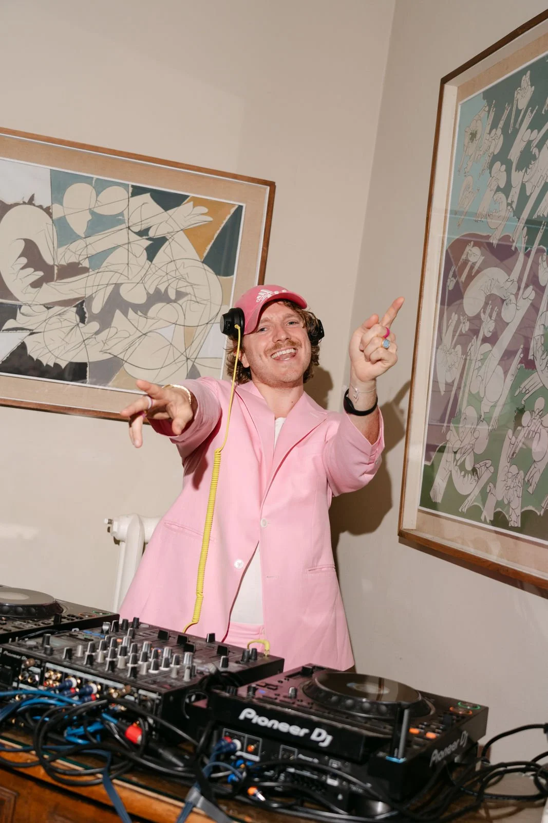 Ein Mann in einem pinken Anzug steht hinter einem DJ-Pult, trägt Kopfhörer und lächelt, während er in die Kamera zeigt. Er befindet sich in einem Raum mit Kunstwerken an den Wänden.