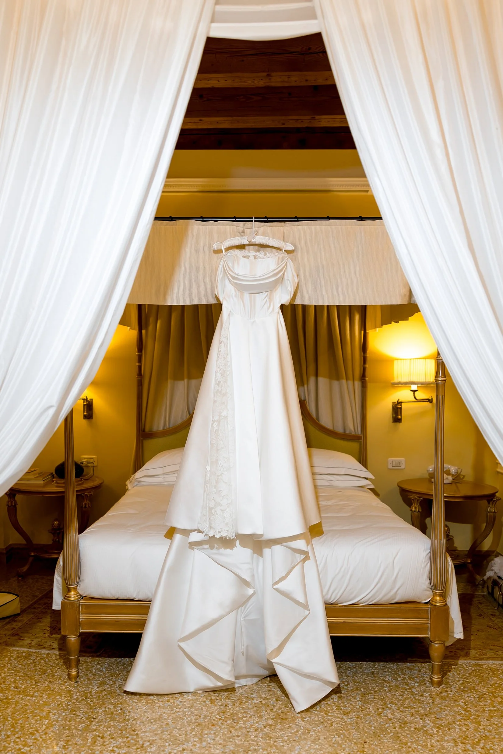 Ein Brautkleid hängt an einem Kleiderbügel über einem Bett in einem Schlafzimmer. Das Bett ist von weißen Vorhängen eingerahmt, die auf beiden Seiten des Bettes hängen, und die Szene ist warm beleuchtet.