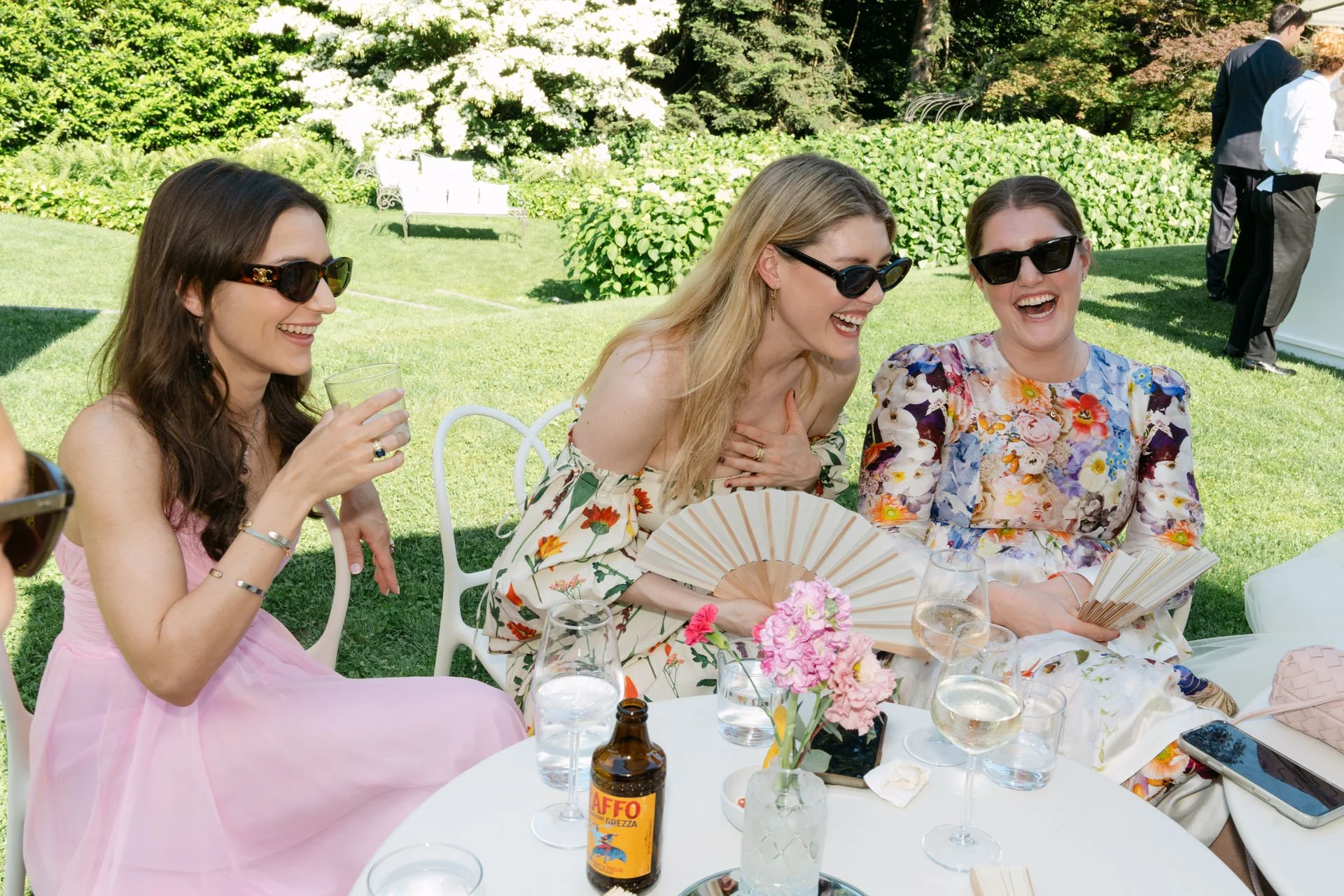 Drei lachende Frauen in eleganten Kleidern sitzen an einem weißen Gartentisch im sonnigen Garten, lachen und unterhalten sich. Im Hintergrund sind grüne Büsche, Bäume und weitere Gäste sichtbar.