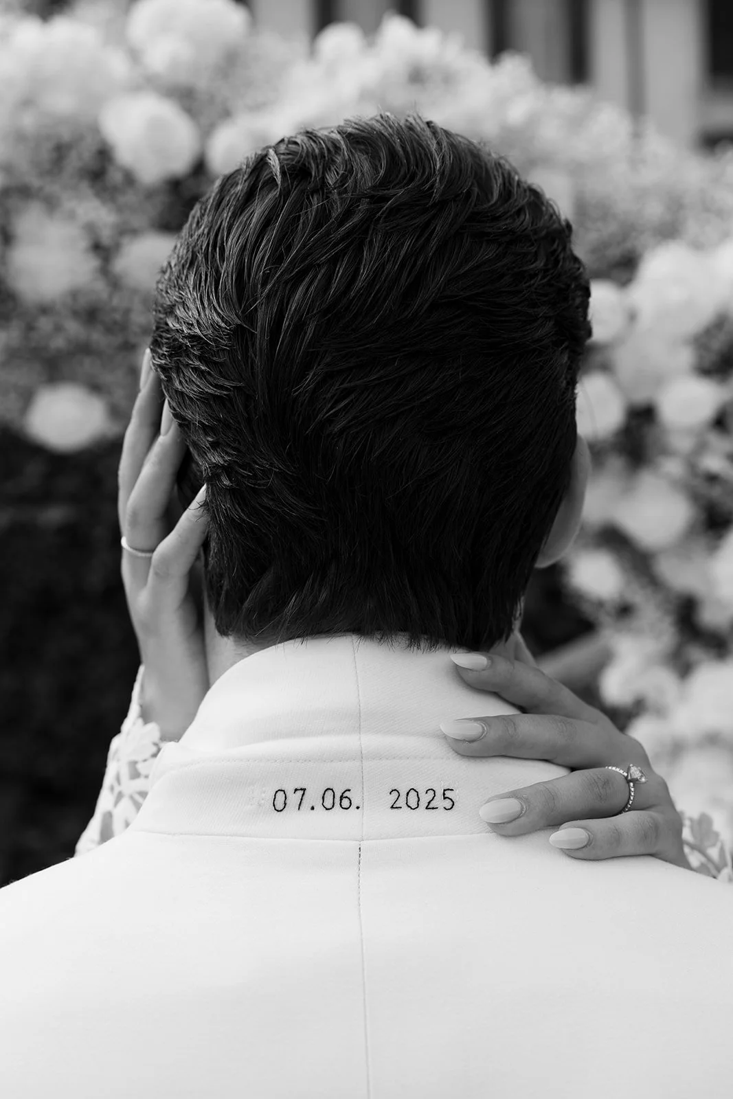 Schwarz-Weiß-Foto einer Person mit kurzen, dunklen Haaren, die den Rücken zeigt. Es trägt ein weißes Kleid oder Hemd mit einem Datum "07.06.2025" auf der Rückseite, während es von zwei Händen gehalten wird. Der Hintergrund besteht aus Blumen.