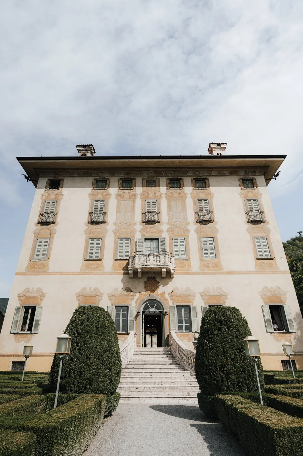 Ein historisches, mehrstöckiges Gebäude mit barocken Elementen, weißen Wänden, mehreren Fenster mit weißen Fensterläden, kleinen Balkonen und einem zentralen Eingang mit Treppe und Gartengestaltung im Vordergrund.